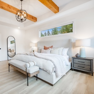 Primary master bedroom with wood beam ceiling hardwood floors white walls sitting area with comfortable chair and books on shelves near staircase with glass walls and wrought iron rails upstairs rooms